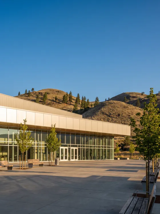 Illustration: a long glass and pale metal arena front with an empty paved plaza, young trees and benches, dry hills with pines behind at golden hour.