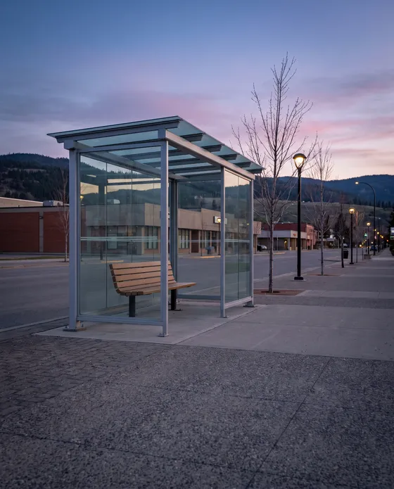 Illustration: an empty glass bus shelter with a bench on a wide street at dusk, street lamps on and hills against a pink sky.