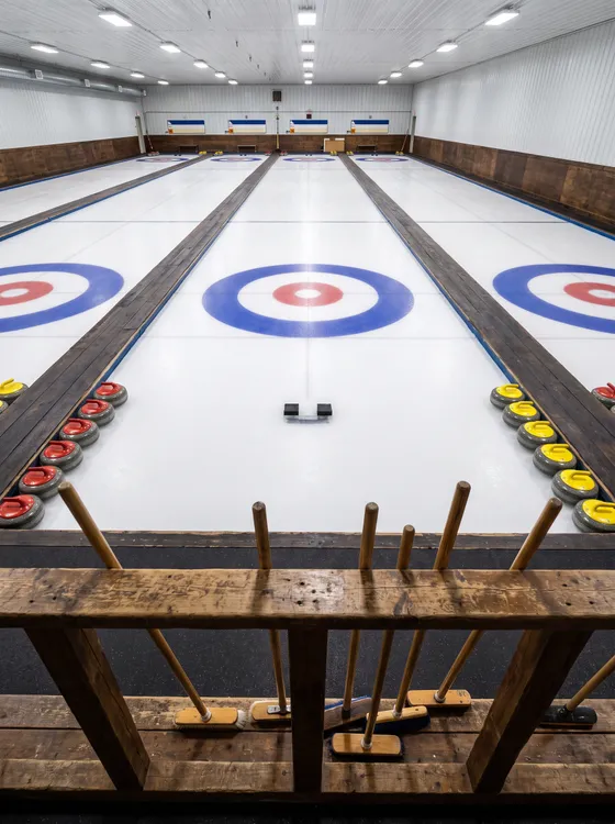 Illustration: an empty curling rink with four white sheets of ice, painted rings and rows of granite stones by the boards.